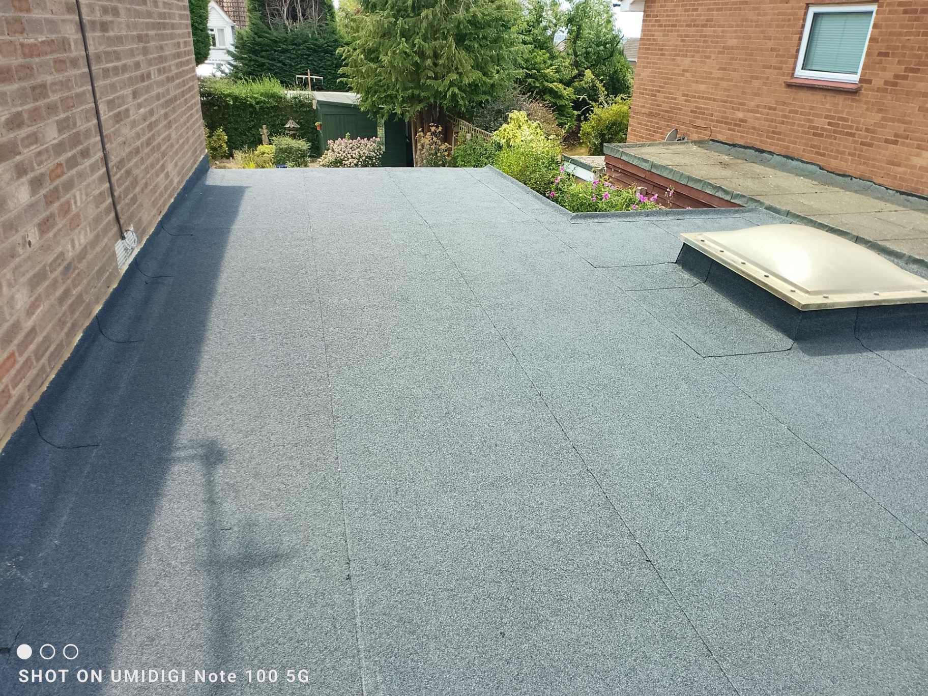 Neatly completed grey torch-on felt flat roof with skylight next to garden
